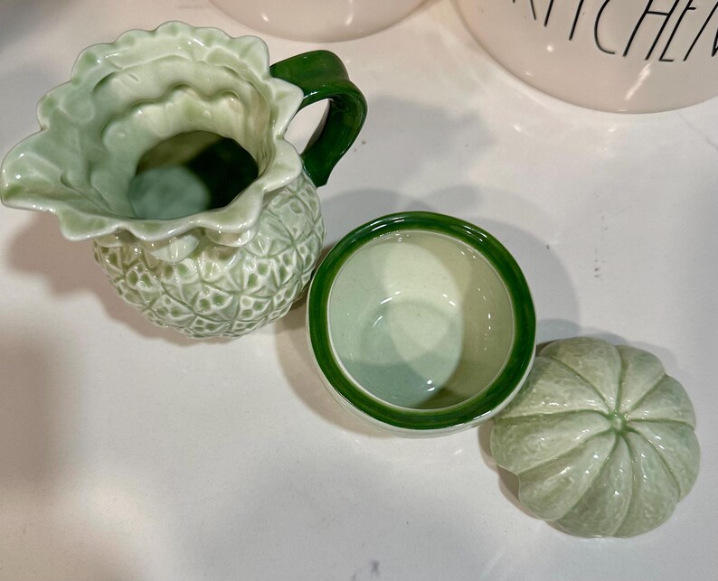 Może przedstawiać: Zestaw trzech zielonych ceramicznych artykuł&oacute;w kuchennych. Artykuły mają kształt ananasa i mają fakturowaną powierzchnię. Zestaw zawiera dzbanek, miskę i pokrywkę.