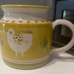 May include: A ceramic mug with a yellow exterior, featuring a white bird illustration with green and orange accents. The mug has a white handle and rim, and green and orange decorative bands. The mug is approximately 4 inches tall.