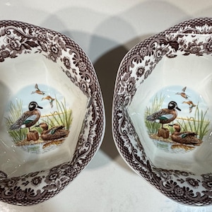 May include: Two white ceramic bowls with a brown floral border and a duck scene in the center. The duck scene features a duck family near water and grass. The bowls have an irregular, scalloped edge.