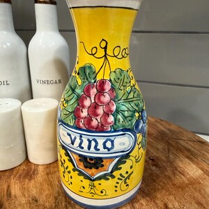 May include: A hand-painted ceramic wine carafe with a yellow body, blue rim, and white accents. The carafe features a grape cluster design with the word "Vino" on a banner. Decorative leaves and vines surround the grapes.
