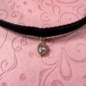 Op de afbeelding: Een zwarte fluwelen choker ketting met een goudkleurige hanger. De hanger heeft een lichtpaarse ovale edelsteen in een goudkleurig, parelrand frame. De achtergrond is een roze oppervlak met een wervelpatroon.