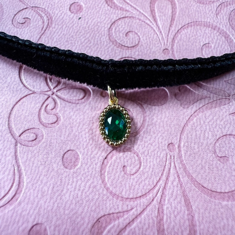 Op de afbeelding: Een zwarte fluwelen choker ketting met een gouden hanger. De hanger heeft een ovale, smaragdgroene edelsteen in een gouden, parelrand. De ketting wordt gepresenteerd op een roze achtergrond.