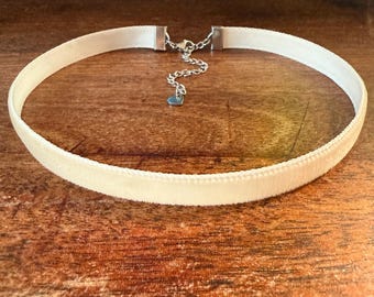 May include: Ivory velvet choker necklace with a silver-toned chain and clasp. The necklace features a small heart-shaped charm and is displayed on a wooden surface.