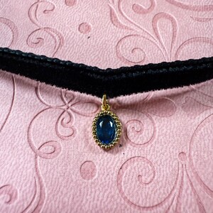 Op de afbeelding: Zwarte fluwelen choker ketting met een gouden hanger. De hanger heeft een grote, ovale, diepblauwe edelsteen in een gouden kralenrand. De achtergrond is een lichtroze, patroon oppervlak.