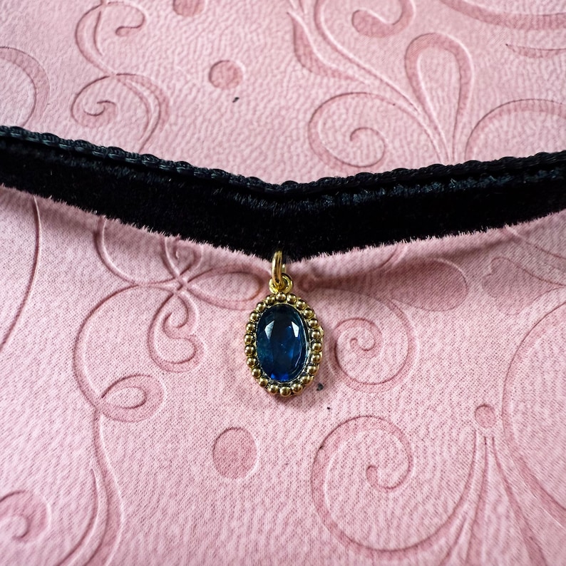 Op de afbeelding: Zwarte fluwelen choker ketting met een gouden hanger. De hanger heeft een grote, ovale, diepblauwe edelsteen in een gouden kralenrand. De achtergrond is een lichtroze, patroon oppervlak.