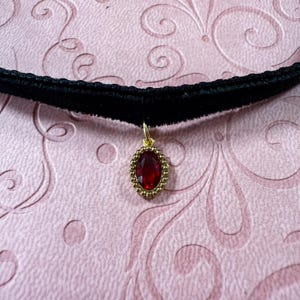 Op de afbeelding: Een zwarte fluwelen choker ketting met een goudkleurige hanger. De hanger heeft een grote, ovale, rode edelsteen omgeven door een kralen gouden rand. De ketting wordt getoond op een roze patroon oppervlak.