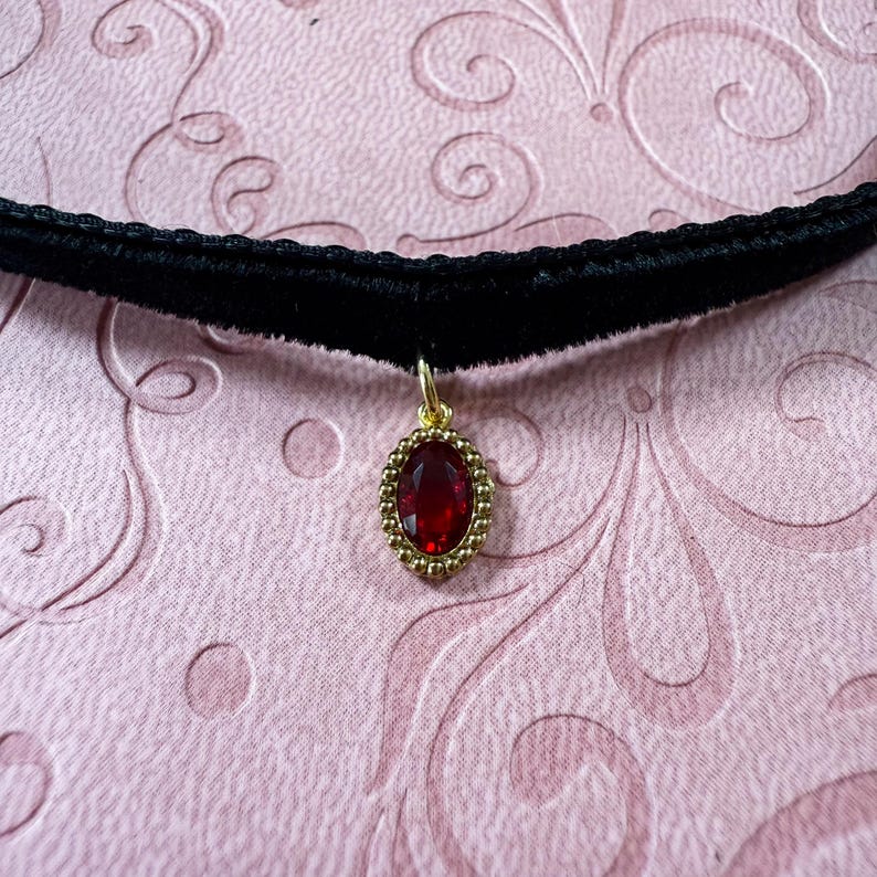Op de afbeelding: Een zwarte fluwelen choker ketting met een goudkleurige hanger. De hanger heeft een grote, ovale, rode edelsteen omgeven door een kralen gouden rand. De ketting wordt getoond op een roze patroon oppervlak.