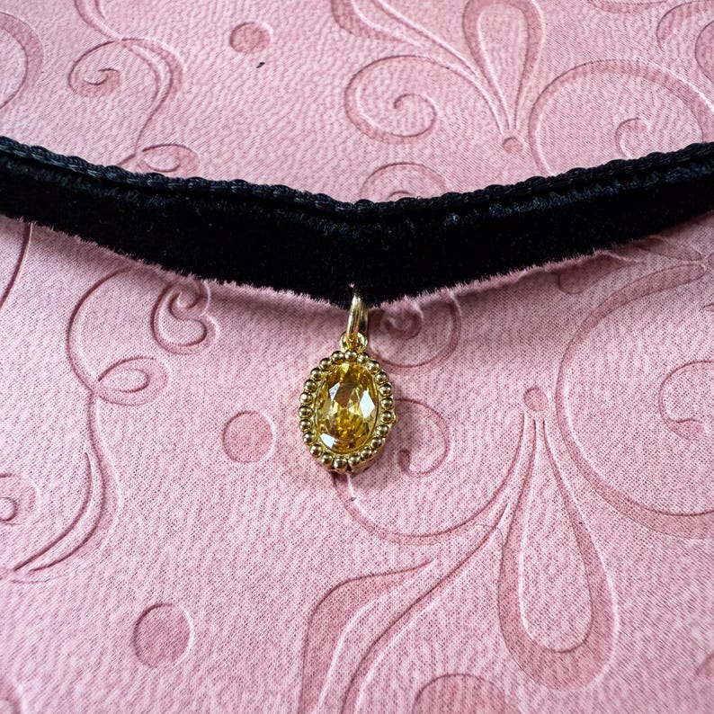 Op de afbeelding: Zwarte fluwelen choker ketting met een gouden hanger. De hanger heeft een ovale, gele edelsteen in een kralen gouden frame. De achtergrond is een roze stof met een wervelend patroon.