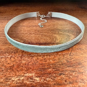 May include: A light blue velvet choker necklace with a silver-toned chain and clasp. The necklace has a small heart-shaped charm. The choker is displayed on a wooden surface.
