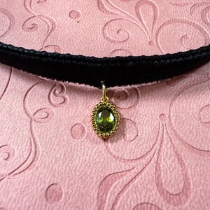 Op de afbeelding: Een zwarte fluwelen choker ketting met een goudkleurige hanger. De hanger heeft een ovale, groene edelsteen met een gouden kralenrand. De ketting wordt getoond op een roze oppervlak met een bloemmotief.
