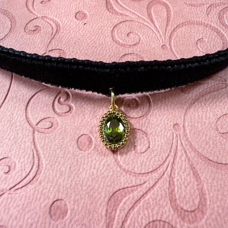 Op de afbeelding: Een zwarte fluwelen choker ketting met een goudkleurige hanger. De hanger heeft een ovale, groene edelsteen met een gouden kralenrand. De ketting wordt getoond op een roze oppervlak met een bloemmotief.