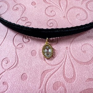 Op de afbeelding: Een zwarte fluwelen choker ketting met een goudkleurige hanger. De hanger heeft een heldere, ovaalvormige steen, omgeven door een kralen gouden frame. De ketting wordt getoond op een roze oppervlak met een wervelend patroon.