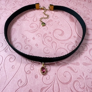 Op de afbeelding: Zwarte fluwelen choker ketting met een goudkleurige sluiting en ketting. De hanger is een ovaalvormige, rode edelsteen met een goudkleurige, kralenrand. De ketting wordt gepresenteerd op een roze achtergrond met een bloemenpatroon.