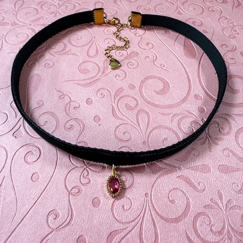 Op de afbeelding: Zwarte fluwelen choker ketting met een goudkleurige sluiting en ketting. De hanger is een ovaalvormige, rode edelsteen met een goudkleurige, kralenrand. De ketting wordt gepresenteerd op een roze achtergrond met een bloemenpatroon.