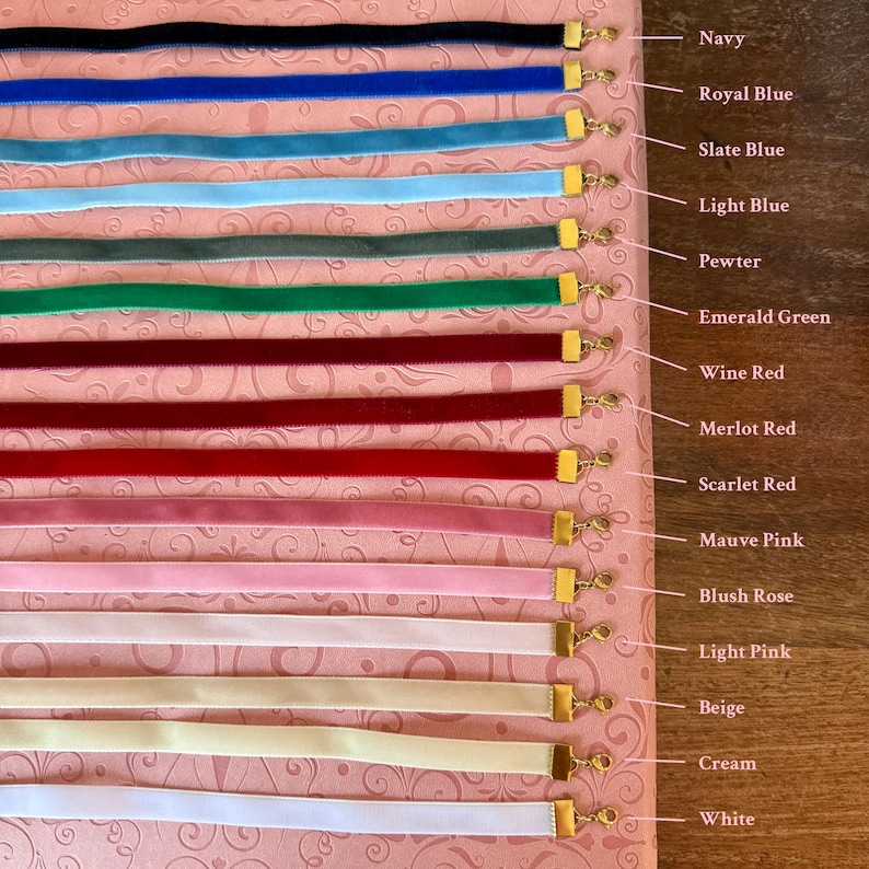 Op de afbeelding: Een verzameling fluwelen lintchokers in verschillende kleuren, waaronder marineblauw, koningsblauw, leisteenblauw, lichtblauw, pewter, smaragdgroen, wijnrood, merlotrood, scharlakenrood, mauve roze, blush roze, lichtroze, beige, cr&egrave;me en wit. Elke choker heeft een goudkleurige sluiting.