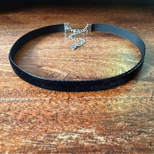 May include: A black velvet choker necklace with a silver-toned chain and clasp. The choker is made of a soft, plush material and is adjustable. The necklace is displayed on a wooden surface.