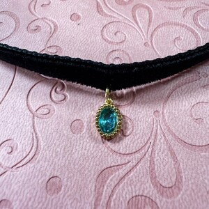 Op de afbeelding: Een zwarte fluwelen choker ketting met een gouden hanger. De hanger heeft een ovale, turquoise edelsteen omgeven door een gouden, kralenomrande lijst. De ketting wordt gepresenteerd op een roze oppervlak met patronen.