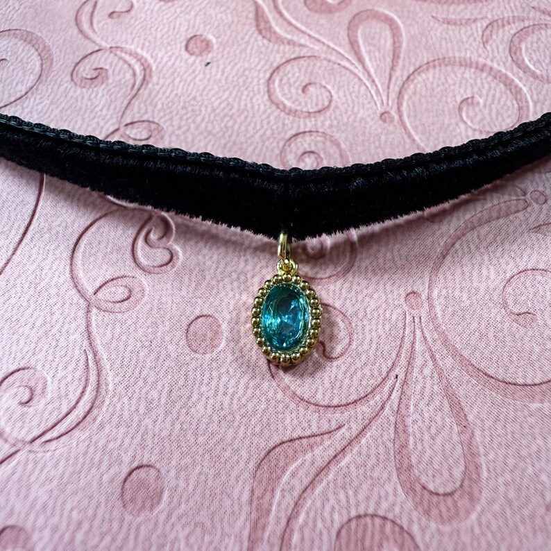 Op de afbeelding: Een zwarte fluwelen choker ketting met een gouden hanger. De hanger heeft een ovale, turquoise edelsteen omgeven door een gouden, kralenomrande lijst. De ketting wordt gepresenteerd op een roze oppervlak met patronen.