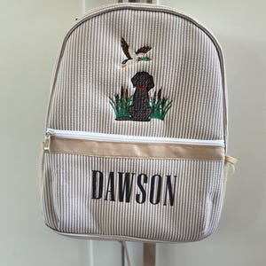 May include: A beige and white striped canvas backpack with a brown dog and a duck embroidered on the front. The backpack has a zippered pocket and a name tag that reads "DAWSON".