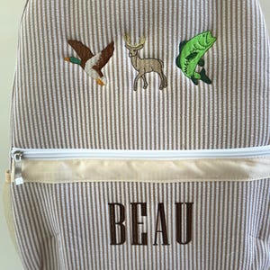 May include: Tan and white striped backpack with embroidered images of a duck, deer, and fish. The name "BEAU" is embroidered in brown letters on the front. Features a white zipper and a small side pocket.