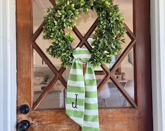 May include: A green wreath with a full, leafy appearance hangs on a wooden door. A green and white striped ribbon with the letter "J" is tied to the wreath. The door has a diamond-shaped window design.