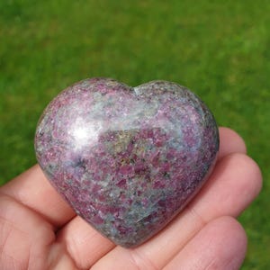 May include: A heart-shaped stone, a mix of pink and grey, with a polished surface. The stone is held in a hand. The background is a blurred green lawn.