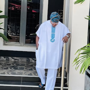 May include: A white tunic and trousers ensemble featuring a blue embroidered detail on the chest. The outfit is complemented by a patterned head wrap and blue sandals. The person is on a set of black and white steps.