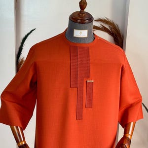 May include: An orange tunic-style shirt with three-quarter sleeves. The shirt features a textured, vertical panel design on the chest and a small rectangular accent. The shirt is displayed on a mannequin.