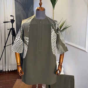 May include: Olive green two-piece outfit with white wave pattern accents on the sleeves. The top has a short zipper and is paired with matching trousers. The outfit is displayed on a wooden mannequin.