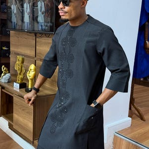 May include: A black, textured, three-quarter sleeve tunic and matching pants. The tunic features an embroidered floral design down the front and a pocket. The outfit is paired with a black cap and sunglasses. The man is wearing a watch and a bracelet.