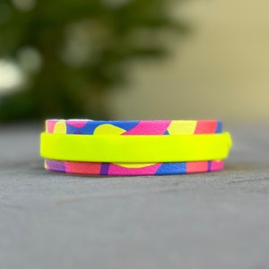Pode incluir: Uma pulseira em camadas com uma faixa central de silicone amarelo néon sólido, ladeada por duas faixas estampadas. As faixas estampadas exibem um design abstrato e colorido com formas giratórias em tons de rosa, azul e amarelo brilhantes.