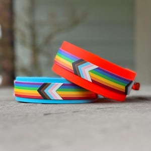 Może przedstawiać: Dwie silikonowe opaski na nadgarstek, jedna w kolorze jaskrawo pomarańczowym, a druga w kolorze jasnoniebieskim, są ułożone jedna na drugiej. Każda opaska ma tęczowy wzór z motywem szewronu, który zawiera kolory flag Pride i Transgender. Opaski leżą na szarej powierzchni.