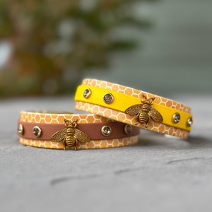 May include: Two bracelets with a honeycomb pattern and a gold bee charm. The bracelets are yellow and brown with clear rhinestones.