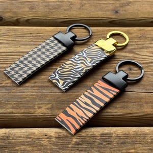 Può includere: Tre portachiavi con diversi motivi su una superficie di legno. Uno ha un motivo a pied-de-poule bianco e nero, un altro un design a strisce zebrate e il terzo presenta strisce di tigre arancioni e nere. Ogni portachiavi ha un anello di metallo.