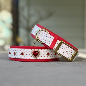 Könnte beinhalten: Zwei rote und weiße Hundehalsbänder mit Herzmuster und goldenen Beschlägen. Das obere Halsband hat einen roten herzförmigen Strassstein in der Mitte.