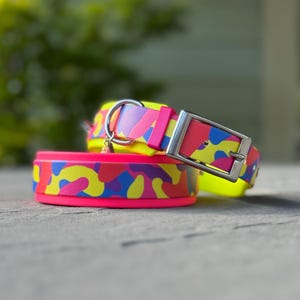 Pode incluir: Uma coleira para animal de estimação com um padrão de camuflagem vibrante em amarelo néon, rosa brilhante e azul. A coleira inclui uma fivela de metal prateado, um anel em D de metal a condizer para a trela e um rebordo rosa néon.