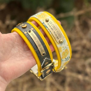 May include: Two dog collars with yellow edges. One is black with gold moon phase icons and a metal tag reading GRIMM 313-686-5555. The other is white with yellow flowers and a metal tag reading ED CINDY YUREK 702 496-4833 | 702 334-5555.