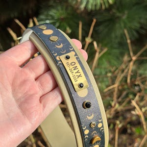 May include: A beige and dark gray pet collar featuring a celestial moon phase pattern in gold. A rectangular brass nameplate is attached, engraved with the text ONYX and the phone number 951-249-5555, accented with two small black circular studs.