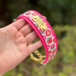 May include: A bright pink pet collar featuring a white and pink gingham pattern with red strawberry illustrations. A rectangular gold-toned metal nameplate is attached, engraved with the text NALA BEAN and a phone number in a clear font.