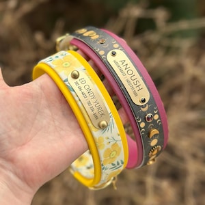 May include: Two pet collars with metal ID tags. One collar is yellow with a yellow floral pattern and a tag reading ED CINDY YUREK 702 496-4833 | 702 334-5555. The other is dark gray with a moon phase pattern and a tag reading ANOUSH (405)8168657 | (405)7955555.