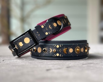Puede incluir: Un collar para mascota de color negro con un patrón celestial de soles, lunas y estrellas doradas. El collar incluye una hebilla negra, una anilla en D y piedras preciosas circulares incrustadas de varios colores, con un forro interior rosa oscuro.
