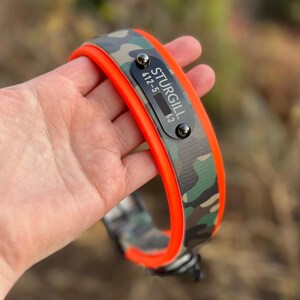 Puede incluir: Un collar con estampado de camuflaje y borde naranja brillante. Una placa de identificaci&oacute;n rectangular negra est&aacute; remachada a la superficie, con el texto blanco STURGILL y el n&uacute;mero 412-555-0163, asegurada con dos remaches de metal oscuro.