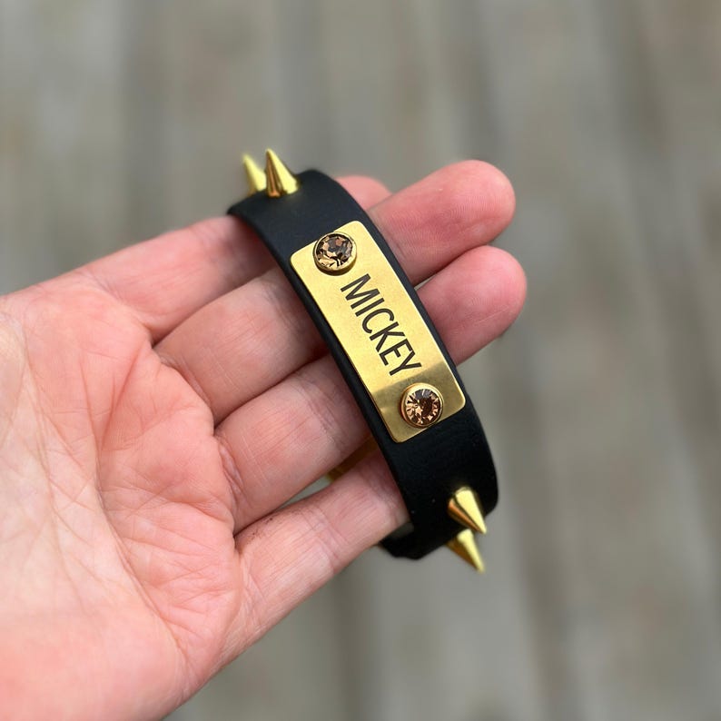 May include: A black band featuring gold-toned metal spikes and a central rectangular gold-toned plate engraved with the name MICKEY. The plate is accented with two round, amber-colored crystals, one positioned above and one below the name.