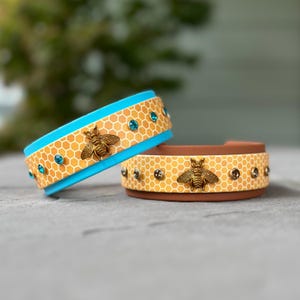 May include: Two cuff bracelets featuring a yellow honeycomb pattern. One bracelet has a bright blue interior and teal crystals, while the other has a brown interior and dark crystals. Each bracelet is adorned with a central brass-toned bee charm.