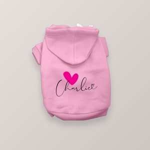 May include: A light pink pet hoodie featuring a hood and short sleeves. The back of the garment displays a bright pink heart graphic positioned above the name Charlie written in a black cursive script font with a small heart at the end.