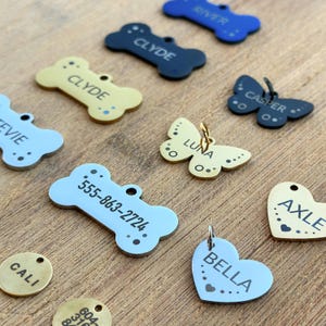 Puede incluir: Surtido de etiquetas de identificaci&oacute;n para mascotas personalizadas en varias formas y colores, incluyendo dise&ntilde;os de hueso, mariposa, coraz&oacute;n y c&iacute;rculo. Las etiquetas presentan nombres grabados como Clyde, Luna, Bella, Axle, Casper y n&uacute;meros de tel&eacute;fono.