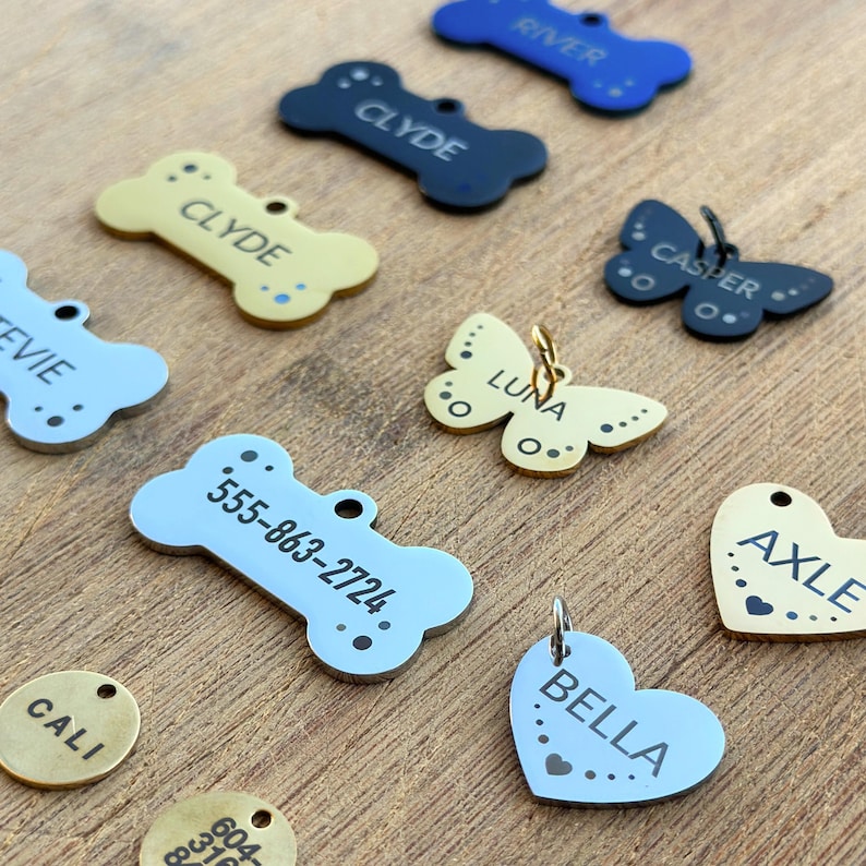 Puede incluir: Surtido de etiquetas de identificaci&oacute;n para mascotas personalizadas en varias formas y colores, incluyendo dise&ntilde;os de hueso, mariposa, coraz&oacute;n y c&iacute;rculo. Las etiquetas presentan nombres grabados como Clyde, Luna, Bella, Axle, Casper y n&uacute;meros de tel&eacute;fono.