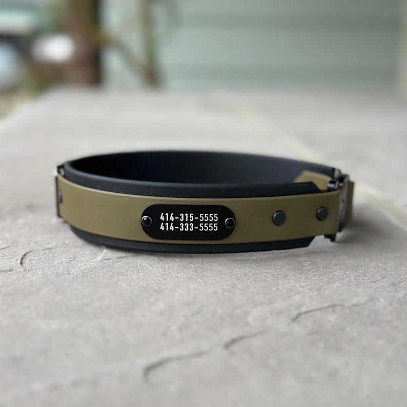 Puede incluir: Un collar de color verde oliva y negro con una placa de identificaci&oacute;n rectangular de metal negro asegurada con dos remaches. La placa muestra los n&uacute;meros de tel&eacute;fono 414-315-5555 y 414-333-5555 impresos en blanco. El collar incluye herrajes negros.
