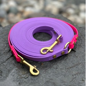 May include: A rolled-up purple dog leash with a bright pink trim and gold-colored metal clasps. The leash is made of a flexible material and is designed for pet owners. The leash is shown on a gray stone surface.