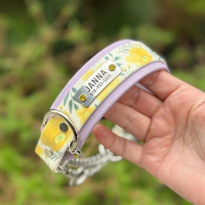 May include: A lavender pet collar featuring a floral pattern with yellow flowers and green leaves on a white background. A rectangular silver metal nameplate is attached, engraved with the name JANNA and the phone number 418-953-5555, accented by two small yellow crystals.